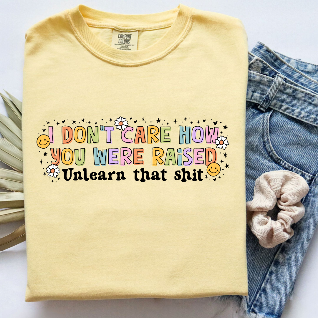 Customized Adult Comfort Colors Butter Yellow Graphic Tee " I don't care how you were raised" rainbow capital letters Unlearn that shit Black bubble letters. Smiley faces and flowers around. good for pride month matching adult crewnecks and hoodies available.