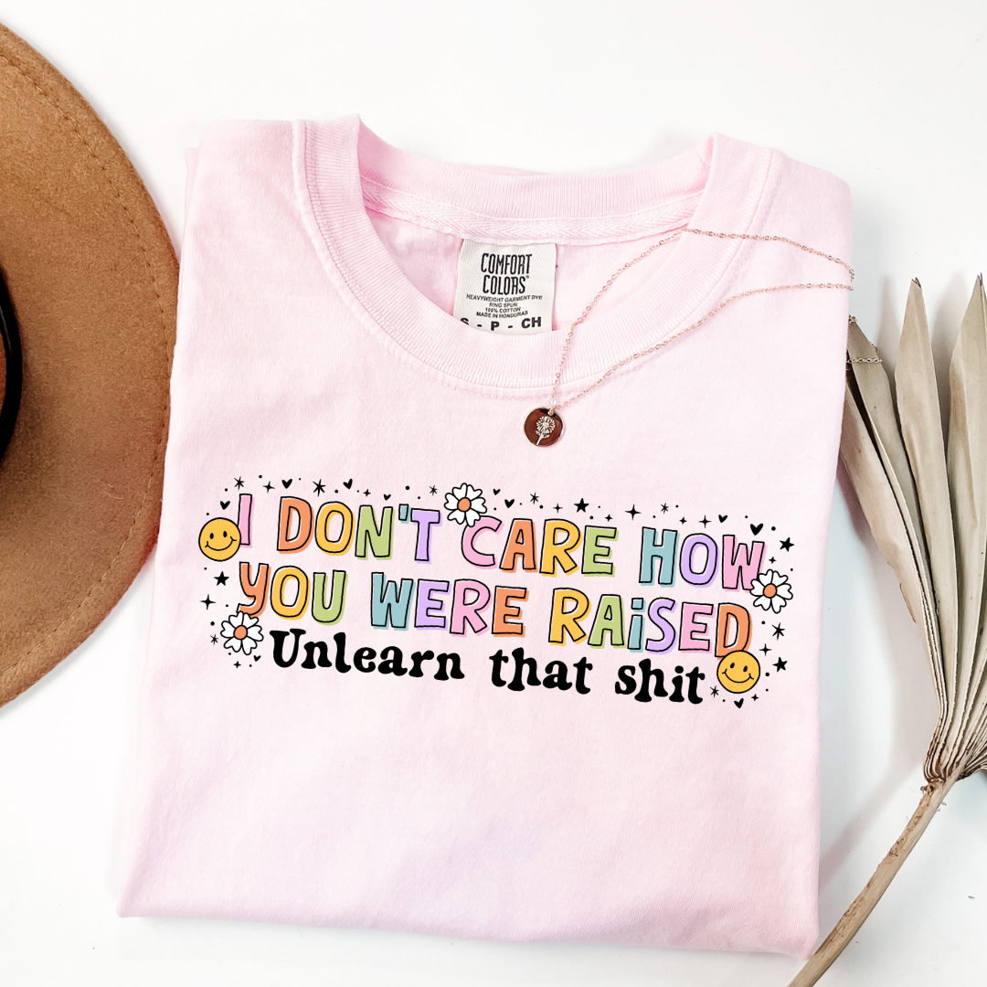 Customized Adult Comfort Colors Blossom Graphic Tee " I don't care how you were raised" rainbow capital letters Unlearn that shit Black bubble letters. Smiley faces and flowers around. good for pride month matching adult crewnecks and hoodies available.