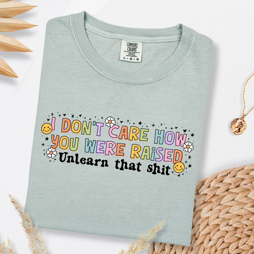 Customized Adult Comfort Colors Bay Graphic Tee " I don't care how you were raised" rainbow capital letters Unlearn that shit Black bubble letters. Smiley faces and flowers around. good for pride month matching adult crewnecks and hoodies available.
