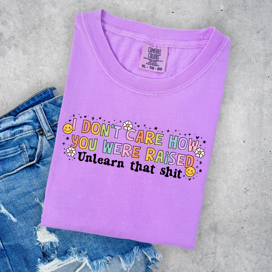 Customized Adult Comfort Colors Neon Violet Graphic Tee " I don't care how you were raised" rainbow capital letters Unlearn that shit Black bubble letters. Smiley faces and flowers around. good for pride month matching adult crewnecks and hoodies available.