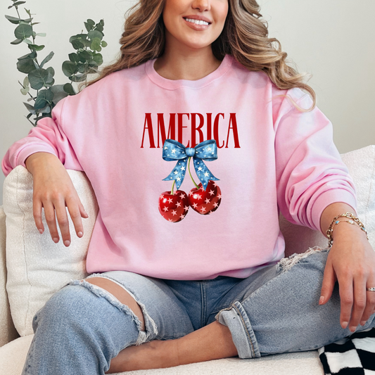 Customized Adult Light Pink Crewneck  Mockup with large VOGUE style Block letters saying America in red with star covered cherries with a Blue star covered Blue Bow, This style is available in adult and Youth Graphic Tees as well as adult crewnecks and hoodies with matching kids Crewnecks. Color availability does very.