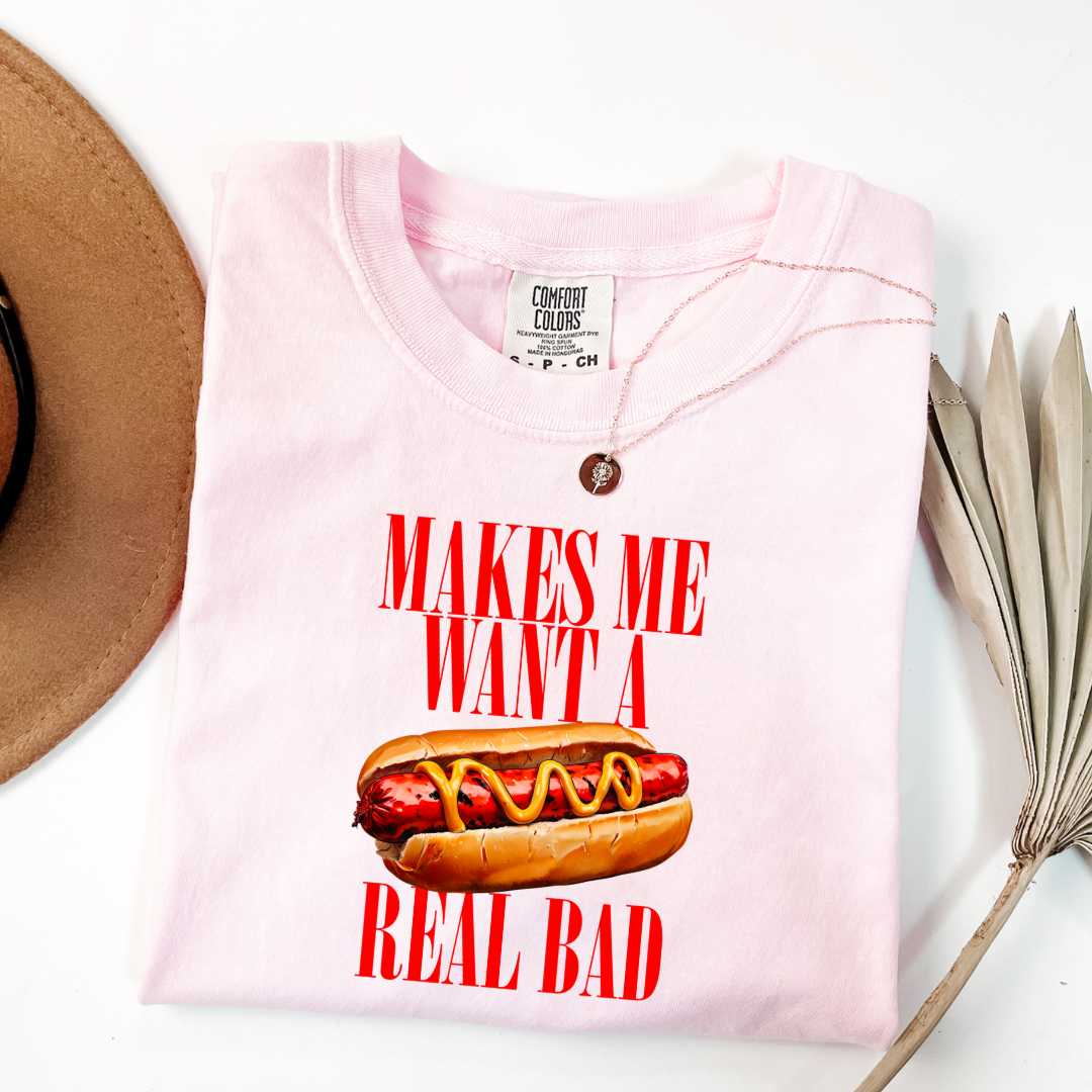 Customized Comfort Colors Blossom Pink Graphic Tee big red block letters " makes me \want a hot dog real bad with a picture of a hotdog in the middle. Jennifer Coolidge quote legally blonde. Available in sizes Youth Small through adult 4X available in matching Crewnecks and adult hoodies as well.