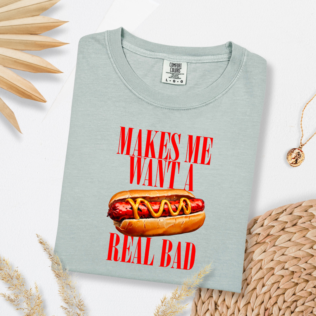 Customized Comfort Colors Bay Green Graphic Tee big red block letters " makes me \want a hot dog real bad with a picture of a hotdog in the middle. Jennifer Coolidge quote legally blonde. Available in sizes Youth Small through adult 4X available in matching Crewnecks and adult hoodies as well.