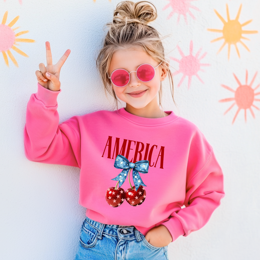 Customized Youth Hot Pink Standing Crewneck  Mockup with large VOGUE style Block letters saying America in red with star covered cherries with a Blue star covered Blue Bow, This style is available in adult and Youth Graphic Tees as well as adult crewnecks and hoodies with matching kids Crewnecks. Color availability does very.
