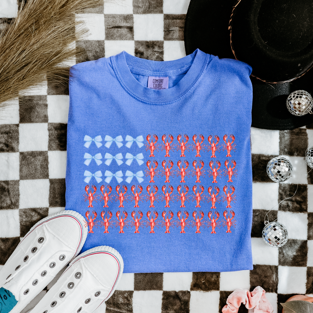 Customized comfort colors Youth Flo Blue Graphic tee with American Flag made of blue bows and red lobsters. Perfect for 4th of July Ranging in sizes youth Small to adult 4X also comes in matching hoodies and crewnecks.