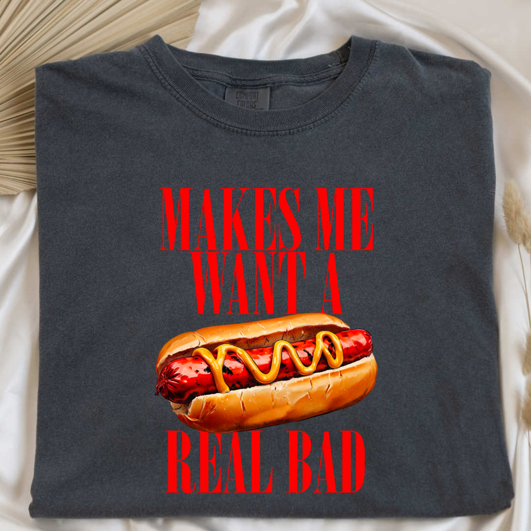 Customized Comfort Colors Pepper Vintage black Graphic Tee big red block letters " makes me \want a hot dog real bad with a picture of a hotdog in the middle. Jennifer Coolidge quote legally blonde. Available in sizes Youth Small through adult 4X available in matching Crewnecks and adult hoodies as well.