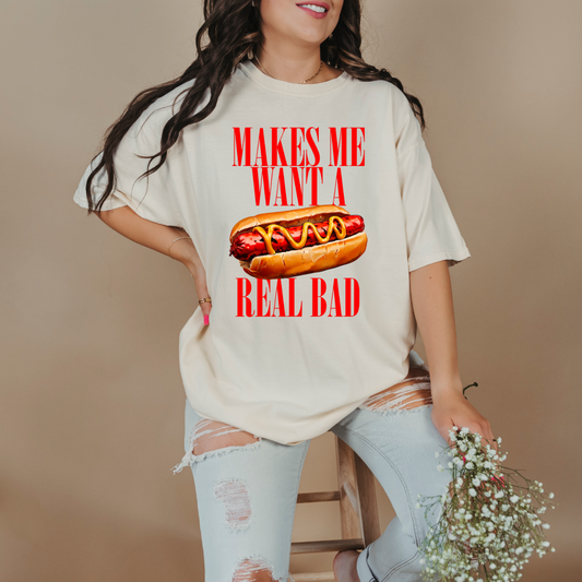 Customized Comfort Colors Ivory Graphic Tee big red block letters " makes me \want a hot dog real bad with a picture of a hotdog in the middle. Jennifer Coolidge quote legally blonde. Available in sizes Youth Small through adult 4X available in matching Crewnecks and adult hoodies as well.