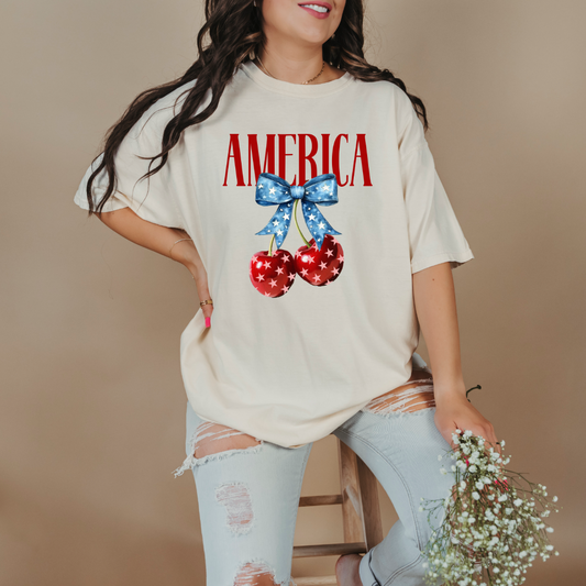Customized Adult Comfort Colors Ivory Standing Mockup with large VOGUE style Block letters saying America in red with star covered cherries with a Blue star covered Blue Bow, This style is available in adult and Youth Graphic Tees as well as adult crewnecks and hoodies with matching kids Crewnecks. Color availability does vary.