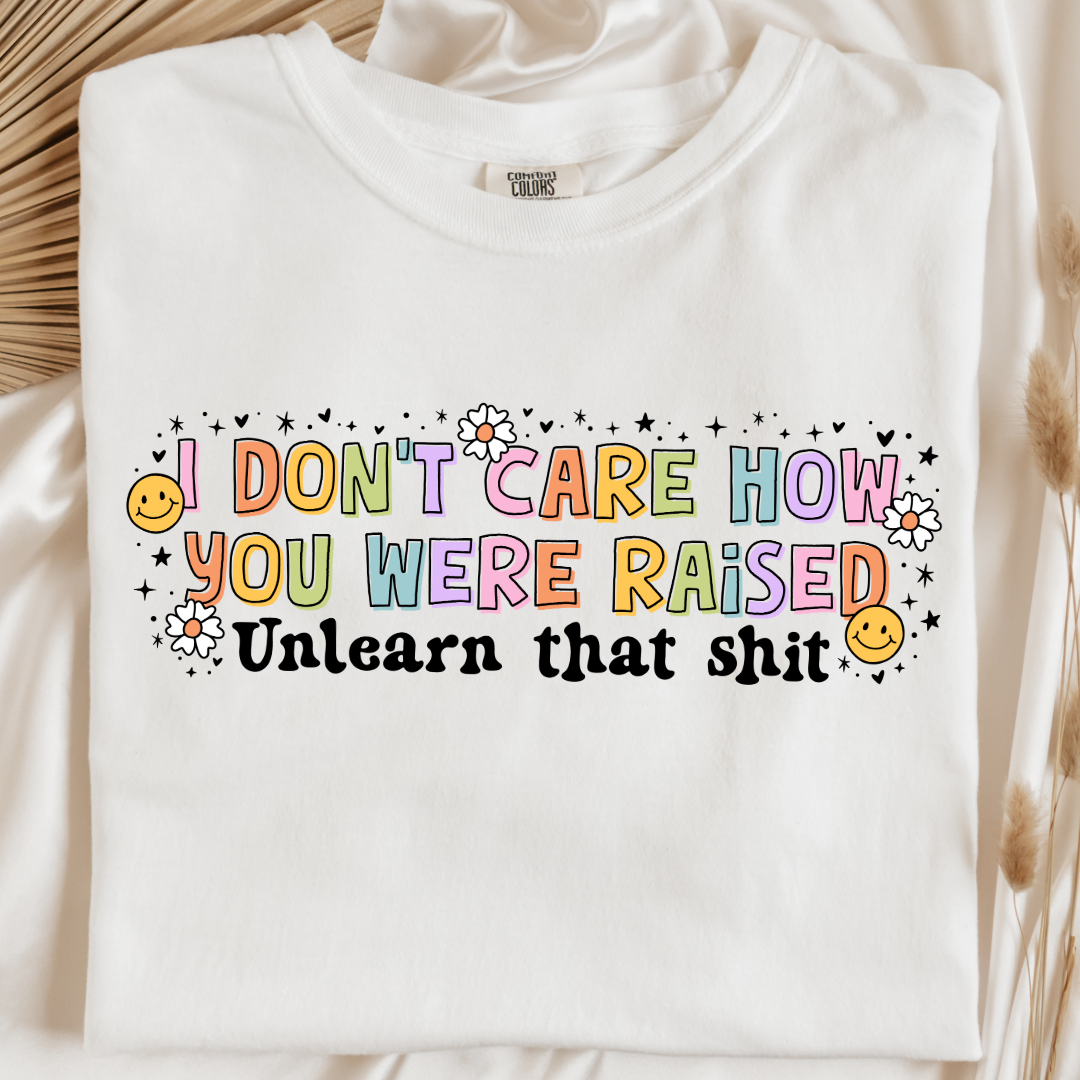 Customized Adult Comfort Colors White Graphic Tee " I don't care how you were raised" rainbow capital letters Unlearn that shit Black bubble letters. Smiley faces and flowers around. good for pride month matching adult crewnecks and hoodies available. 