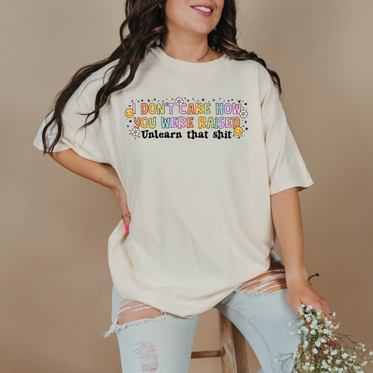 Customized Adult Comfort Colors Ivory Graphic Tee " I don't care how you were raised" rainbow capital letters Unlearn that shit Black bubble letters. Smiley faces and flowers around. good for pride month matching adult crewnecks and hoodies available. 