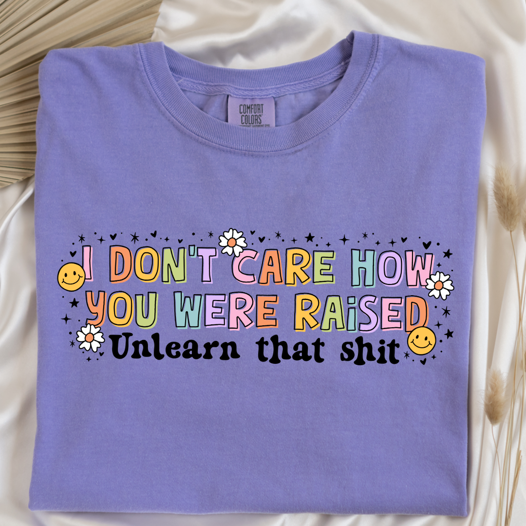 Customized Adult Comfort Colors Violet Graphic Tee " I don't care how you were raised" rainbow capital letters Unlearn that shit Black bubble letters. Smiley faces and flowers around. good for pride month matching adult crewnecks and hoodies available. 