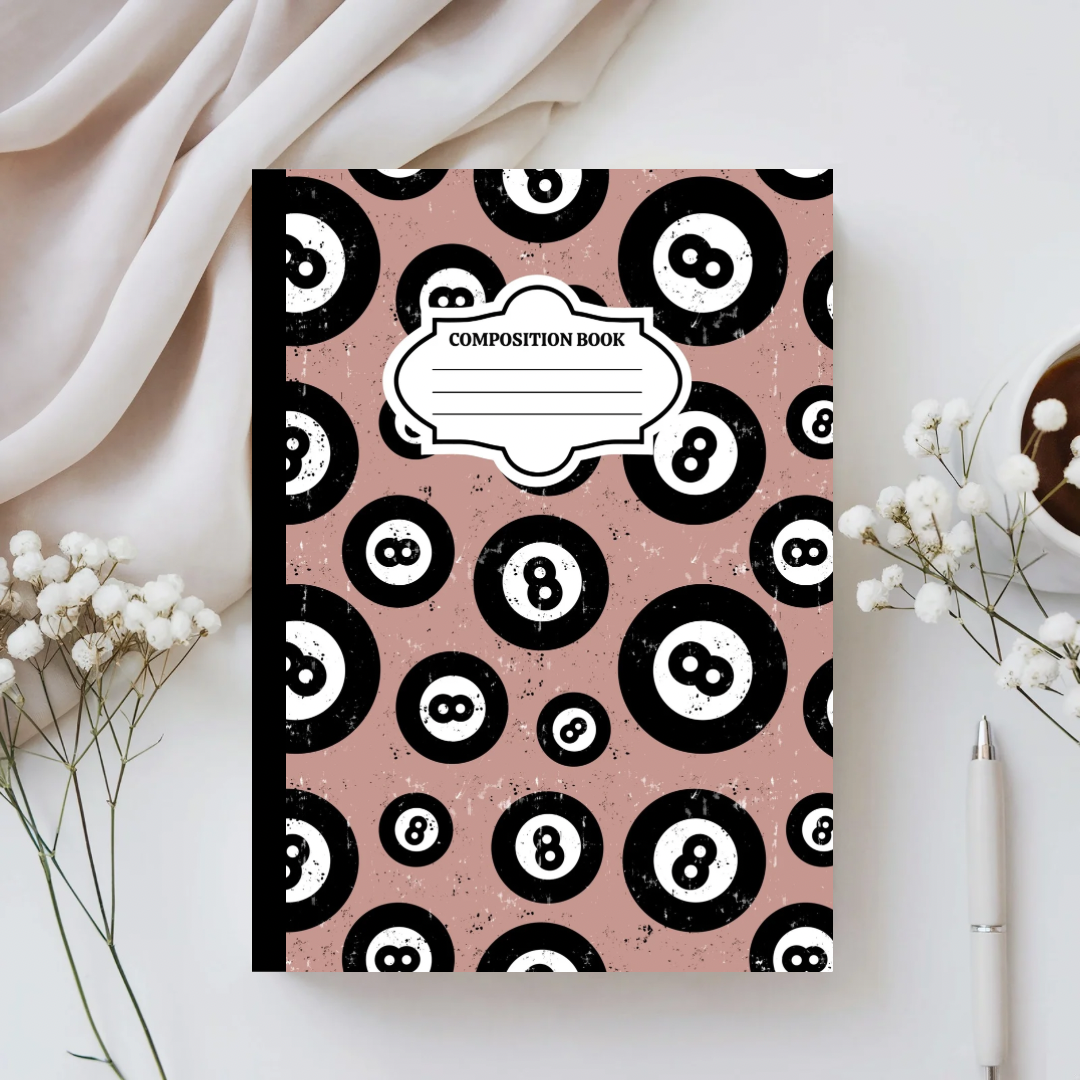 Composition book with black and white eight-ball pattern on a pink background, placed on a light surface with decorative elements.