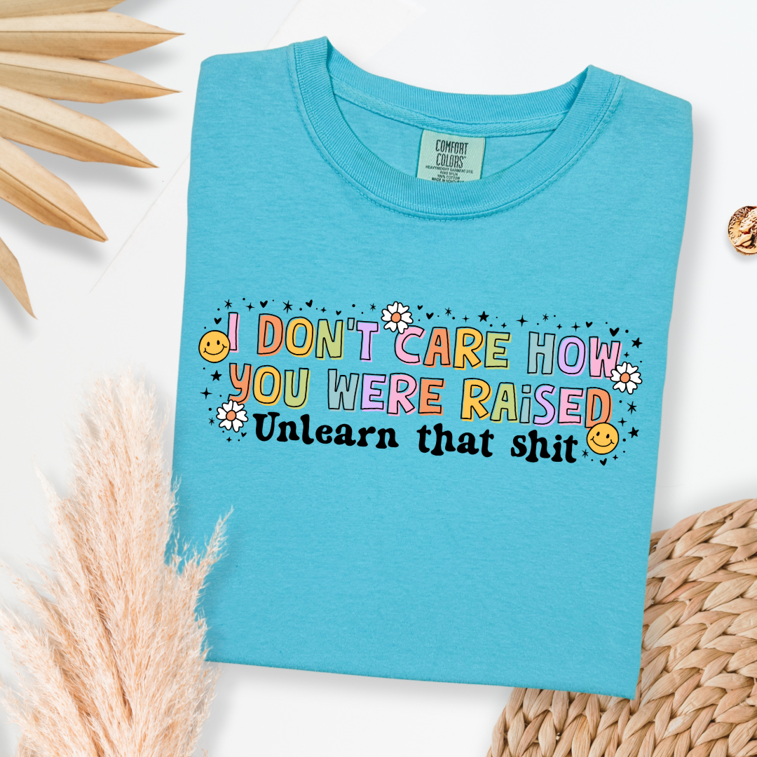 Customized Adult Comfort Colors Lagoon Blue Graphic Tee " I don't care how you were raised" rainbow capital letters Unlearn that shit Black bubble letters. Smiley faces and flowers around. good for pride month matching adult crewnecks and hoodies available. 