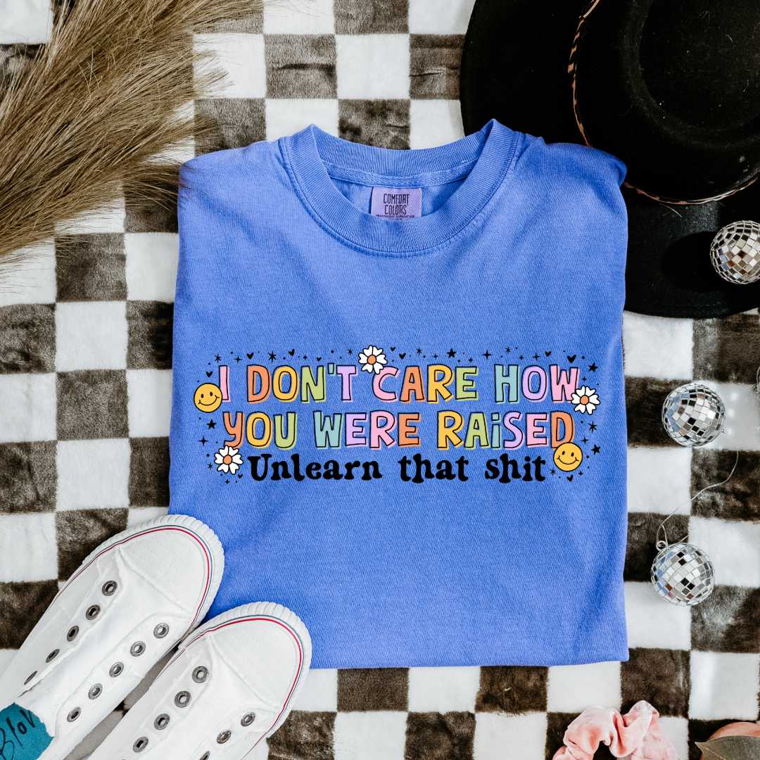 Customized Adult Comfort Colors Flo Blue Graphic Tee " I don't care how you were raised" rainbow capital letters Unlearn that shit Black bubble letters. Smiley faces and flowers around. 