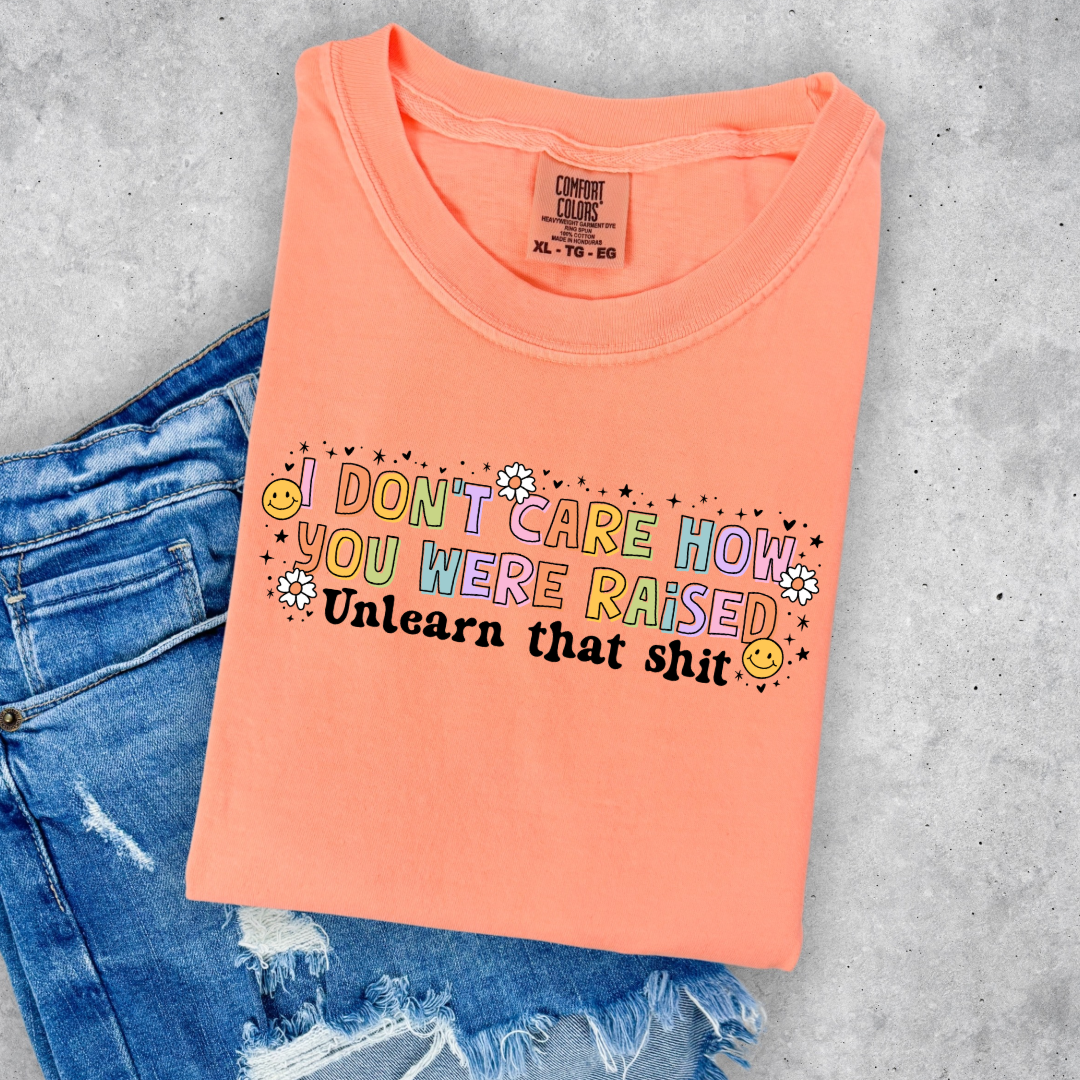 Customized Adult Comfort Colors Neon Cantaloupe Graphic Tee " I don't care how you were raised" rainbow capital letters Unlearn that shit Black bubble letters. Smiley faces and flowers around. good for pride month matching adult crewnecks and hoodies available. 