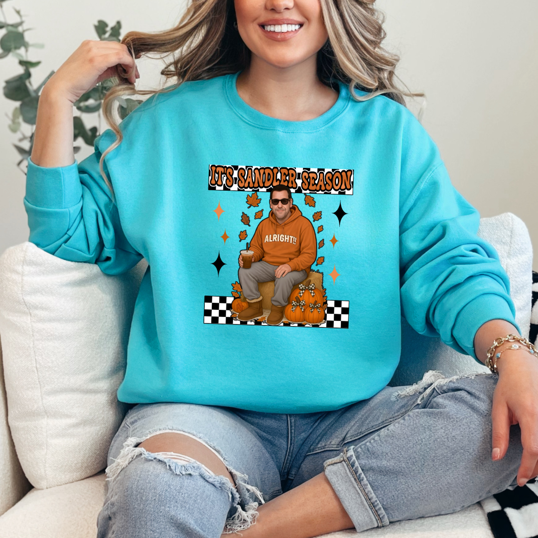 Lagoon blue Adult Crewneck with Adam Sandler on the front sitting on a hay bale stack with a coffee in hand wearing UGGS and checker board its Sandler season. Design available in graphic tees as well in Adult sizing ranging from Small to 4X.