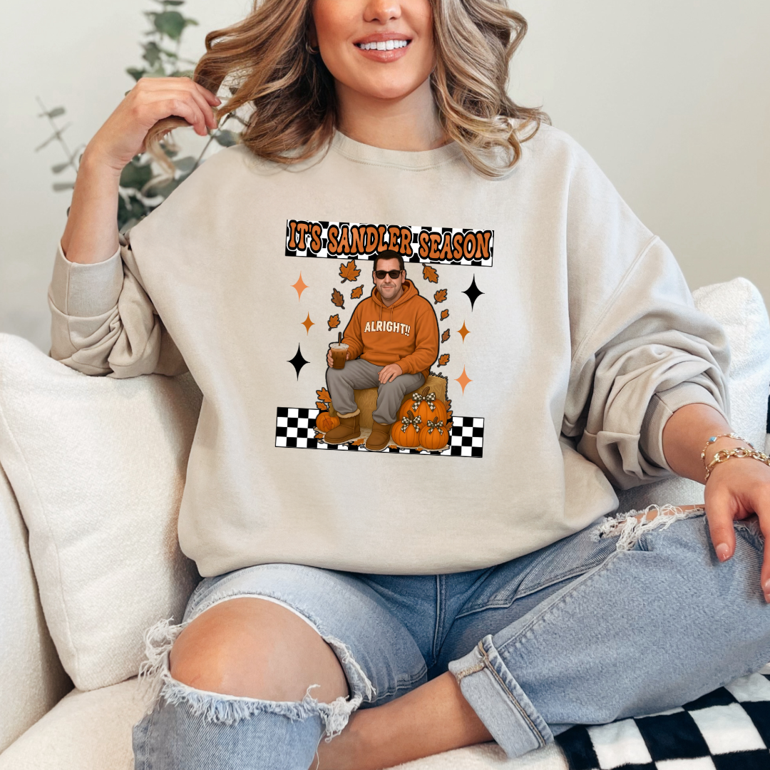 Beige Adult Crewneck with Adam Sandler on the front sitting on a hay bale stack with a coffee in hand wearing UGGS and checker board its Sandler season. Design available in graphic tees as well in Adult sizing ranging from Small to 4X.