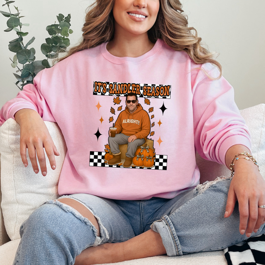 Light Pink Adult Crewneck with Adam Sandler on the front sitting on a hay bale stack with a coffee in hand wearing UGGS and checker board its Sandler season. Design available in graphic tees as well in Adult sizing ranging from Small to 4X.