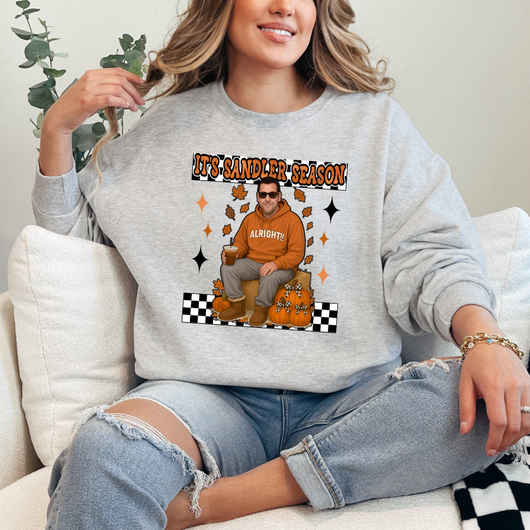 Ash Grey Adult Crewneck with Adam Sandler on the front sitting on a hay bale stack with a coffee in hand wearing UGGS and checker board its Sandler season. Design available in graphic tees as well in Adult sizing ranging from Small to 4X.