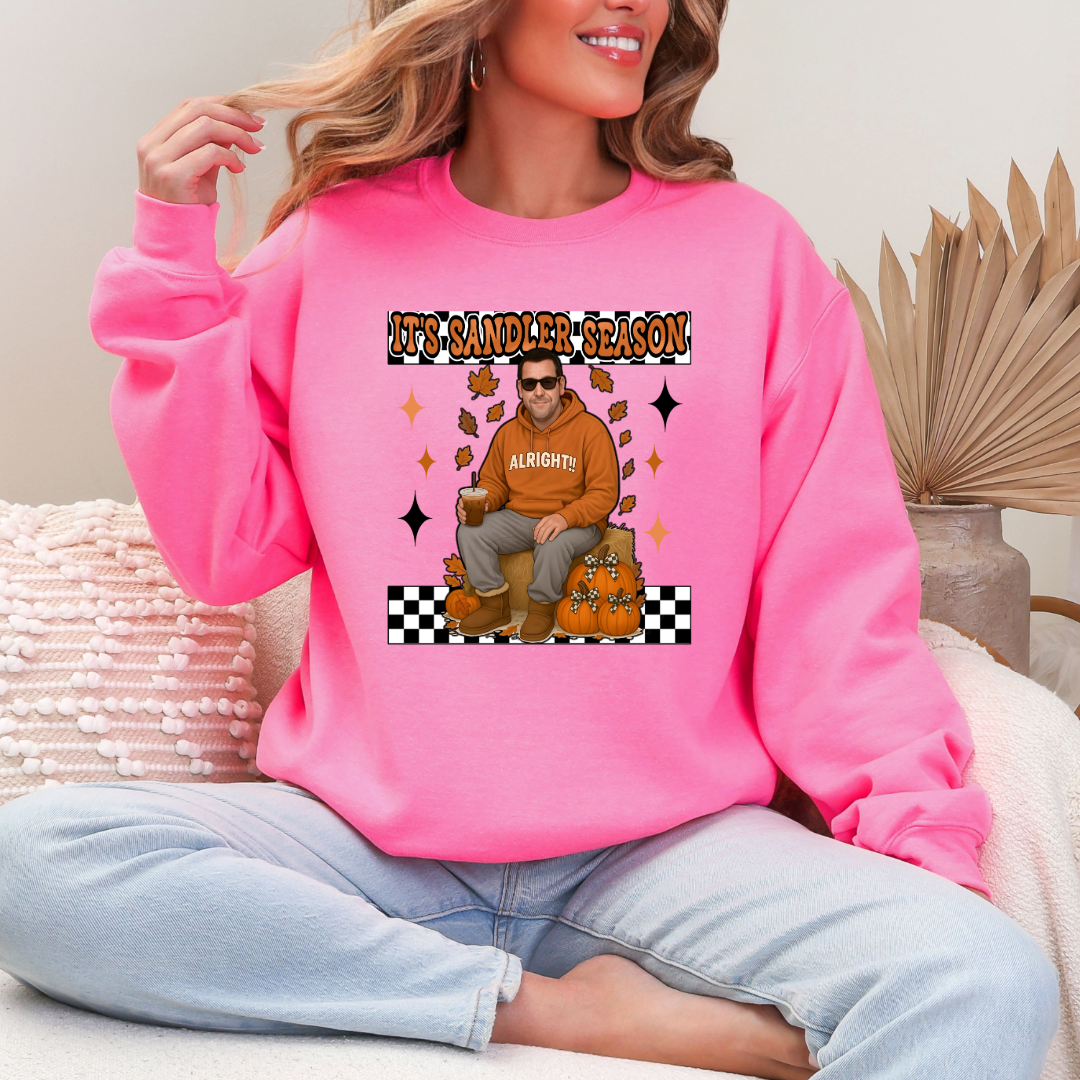 Hot Pink Adult Crewneck with Adam Sandler on the front sitting on a hay bale stack with a coffee in hand wearing UGGS and checker board its Sandler season. Design available in graphic tees as well in Adult sizing ranging from Small to 4X.