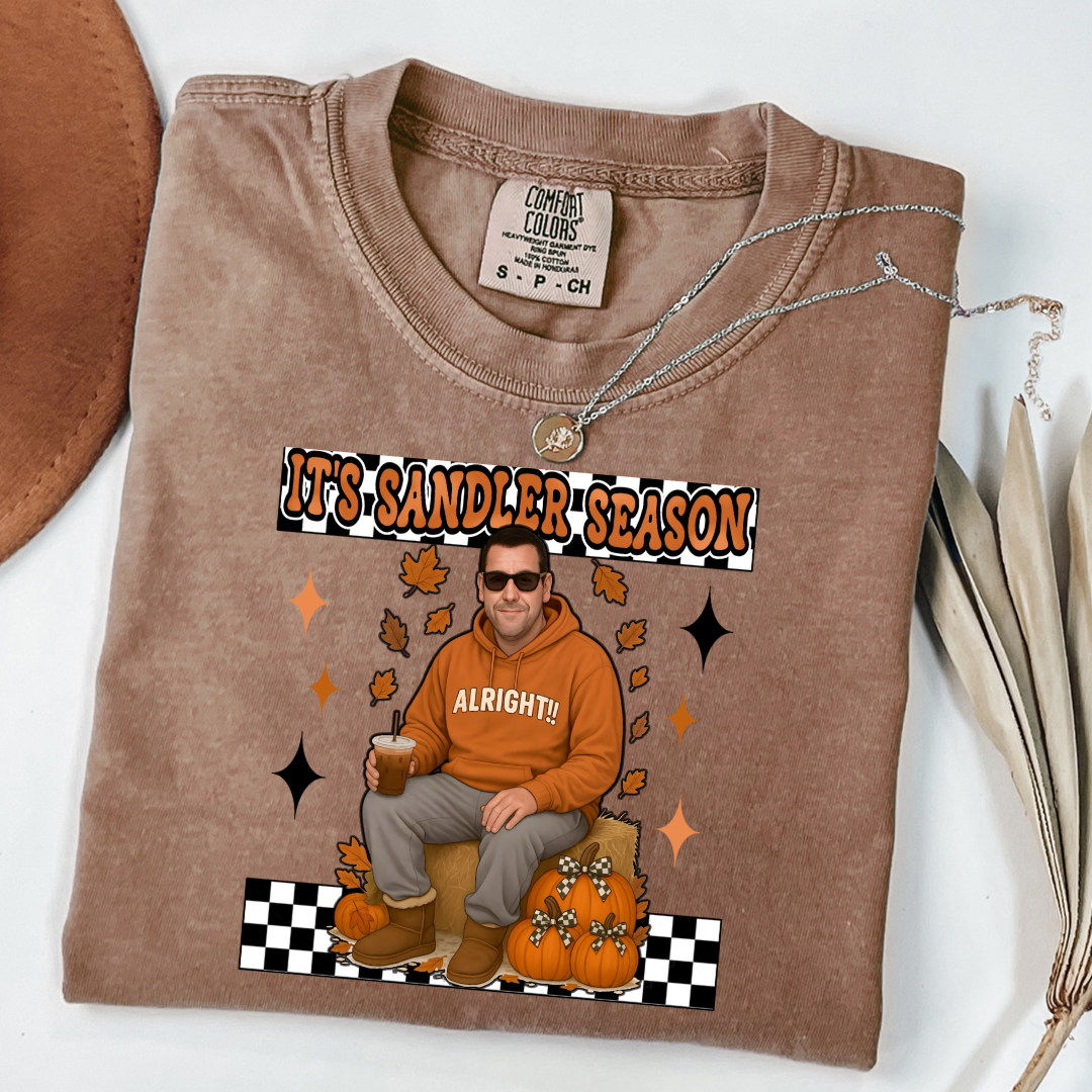 Comfort Colors Espresso Brown Adult Graphic Tee with Adam Sandler on the front sitting on a hay bale stack with a coffee in hand wearing UGGS and checker board its Sandler season. Design available in graphic tees as well in Adult sizing ranging from Small to 4X.