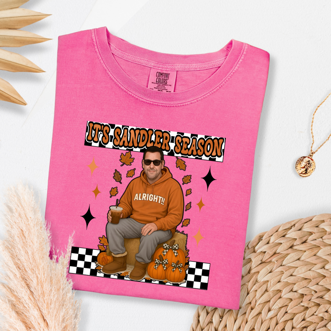 Comfort Colors Crunchberry Pink Adult Graphic Tee with Adam Sandler on the front sitting on a hay bale stack with a coffee in hand wearing UGGS and checker board its Sandler season. Design available in graphic tees as well in Adult sizing ranging from Small to 4X.