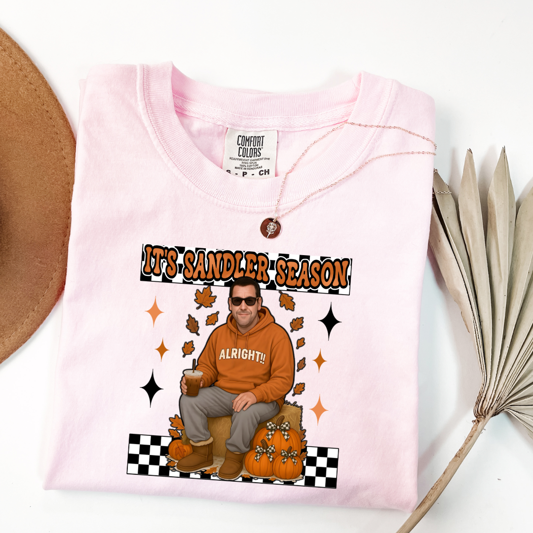 Comfort Colors Blossom Pink Adult Graphic Tee with Adam Sandler on the front sitting on a hay bale stack with a coffee in hand wearing UGGS and checker board its Sandler season. Design available in graphic tees as well in Adult sizing ranging from Small to 4X.