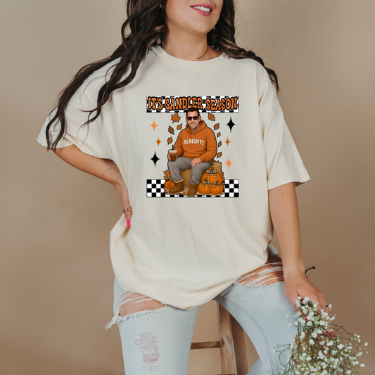 Comfort Colors Ivory Adult Graphic Tee with Adam Sandler on the front sitting on a hay bale stack with a coffee in hand wearing UGGS and checker board its Sandler season. Design available in graphic tees as well in Adult sizing ranging from Small to 4X.