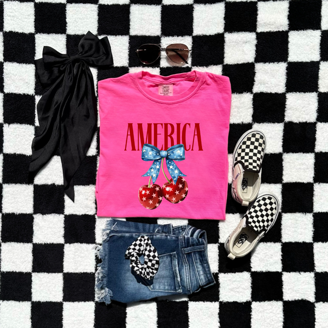 Customized Youth  Comfort Colors Neon Pink  Mockup with large VOGUE style Block letters saying America in red with star covered cherries with a Blue star covered Blue Bow, This style is available in adult and Youth Graphic Tees as well as adult crewnecks and hoodies with matching kids Crewnecks. Color availability does very. 