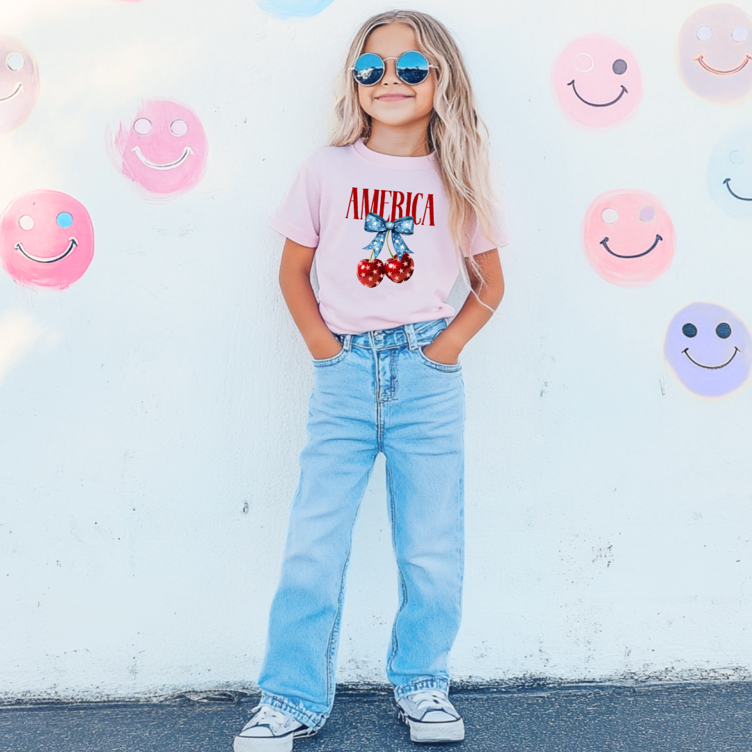Customized Youth  Comfort Colors Standing Blossom Pink  Mockup with large VOGUE style Block letters saying America in red with star covered cherries with a Blue star covered Blue Bow, This style is available in adult and Youth Graphic Tees as well as adult crewnecks and hoodies with matching kids Crewnecks. Color availability does very. 