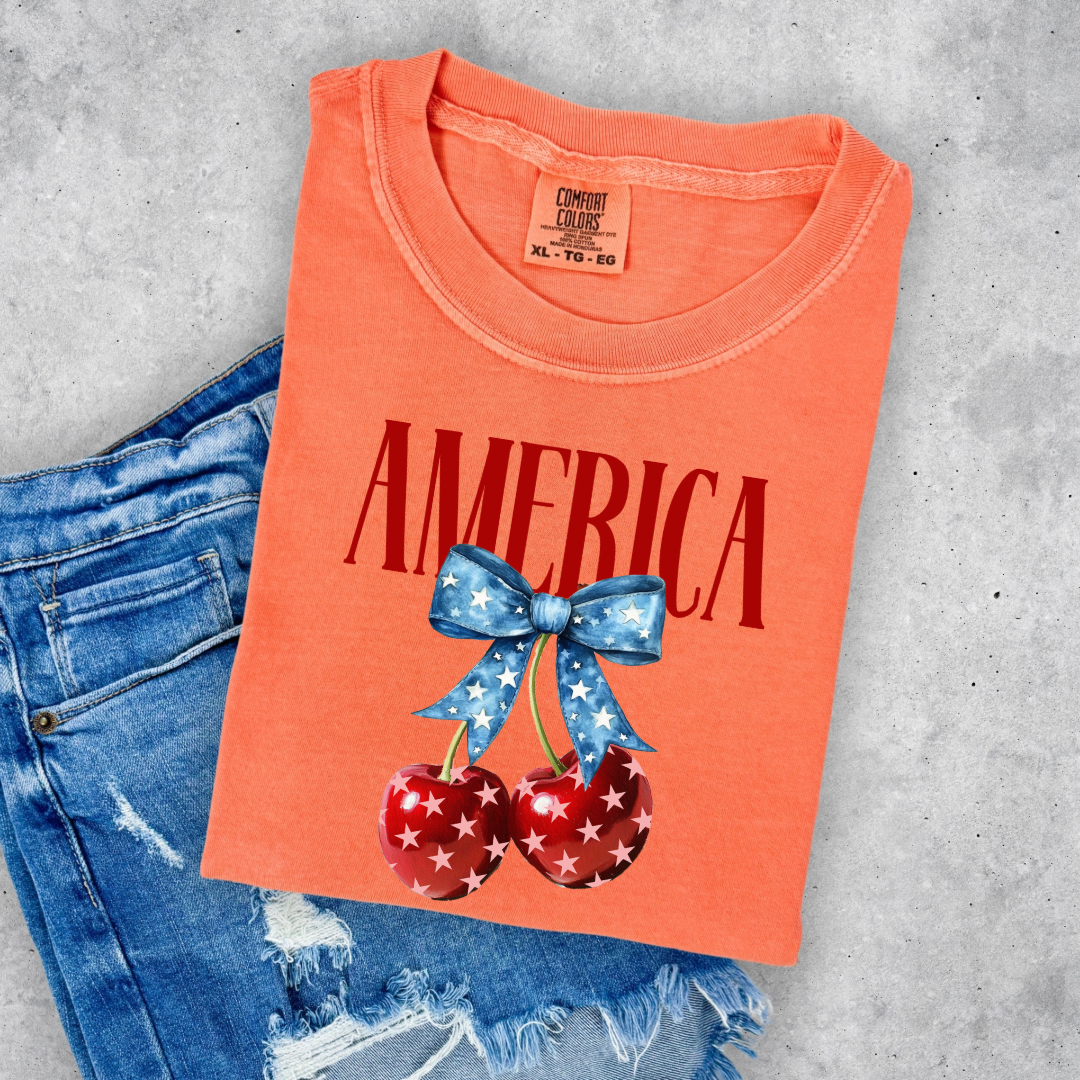 Customized Youth  Comfort Colors Melon Orange Mockup with large VOGUE style Block letters saying America in red with star covered cherries with a Blue star covered Blue Bow, This style is available in adult and Youth Graphic Tees as well as adult crewnecks and hoodies with matching kids Crewnecks. Color availability does very. 