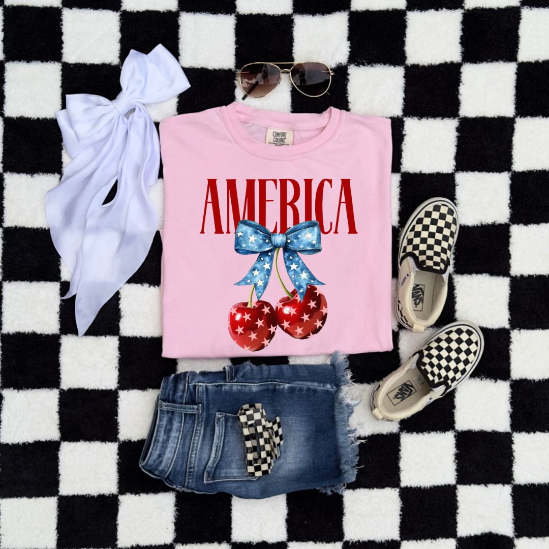 Customized Youth  Comfort Colors Folded Blossom Pink  Mockup with large VOGUE style Block letters saying America in red with star covered cherries with a Blue star covered Blue Bow, This style is available in adult and Youth Graphic Tees as well as adult crewnecks and hoodies with matching kids Crewnecks. Color availability does very. 