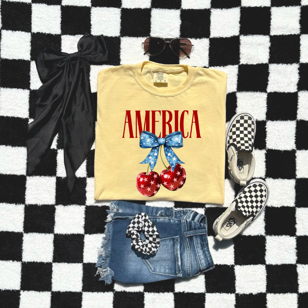 Customized Youth  Comfort Colors Standing Butter Yellow Mockup with large VOGUE style Block letters saying America in red with star covered cherries with a Blue star covered Blue Bow, This style is available in adult and Youth Graphic Tees as well as adult crewnecks and hoodies with matching kids Crewnecks. Color availability does very. 